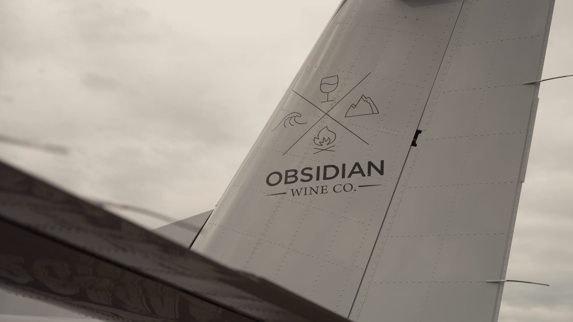 Close-up of the tail of a small aircraft marked 'Obsidian Wine Co.' with wave, mountain, and flame icons