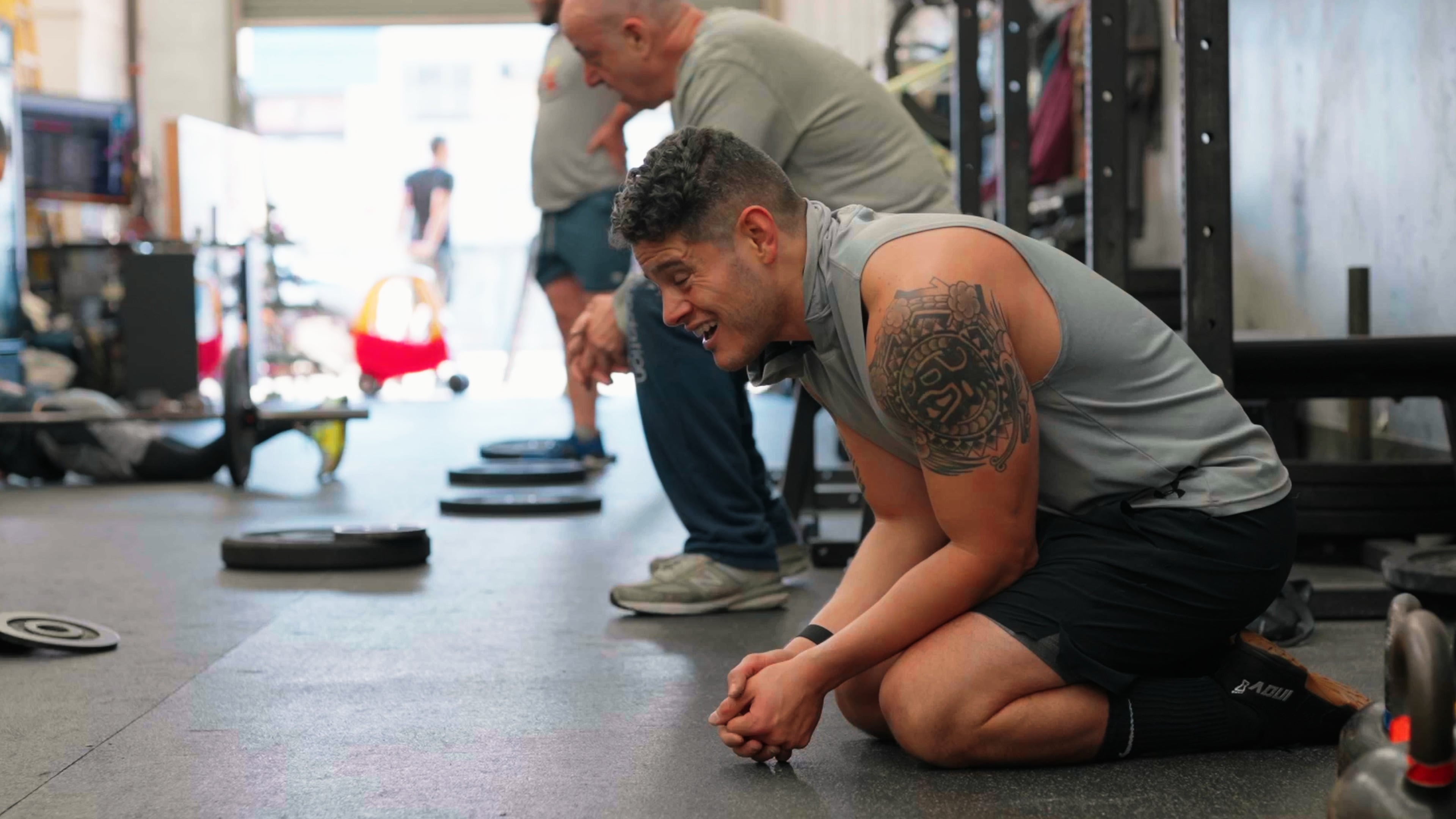 Member kneeling on the gym floor laughing after a workout, others training behind him