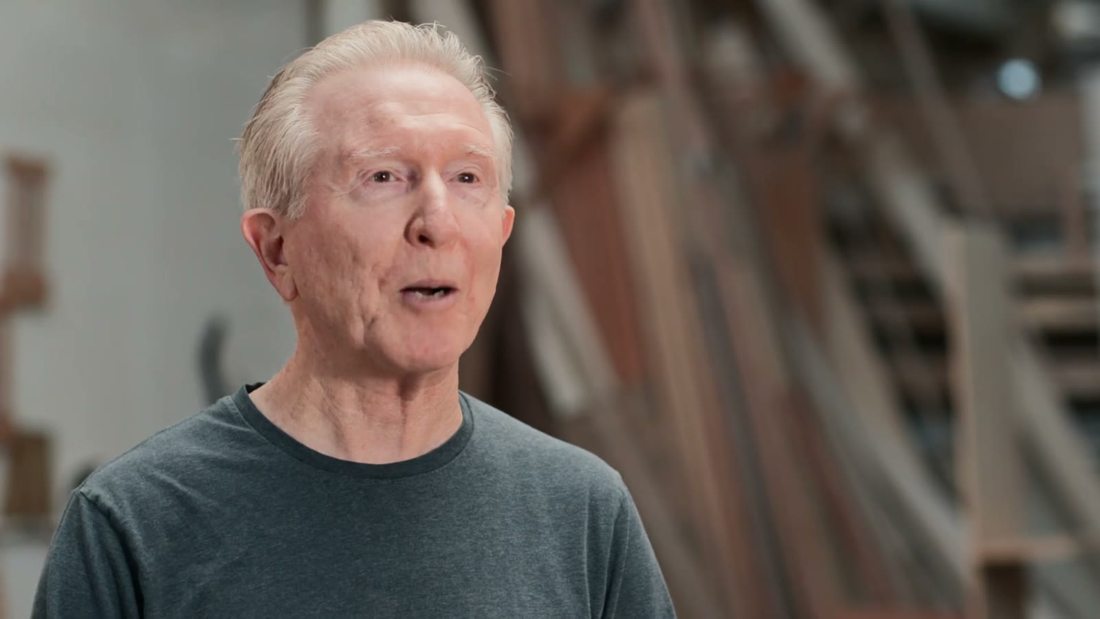 Older man speaking to camera in a woodworking workshop, boards stacked behind him